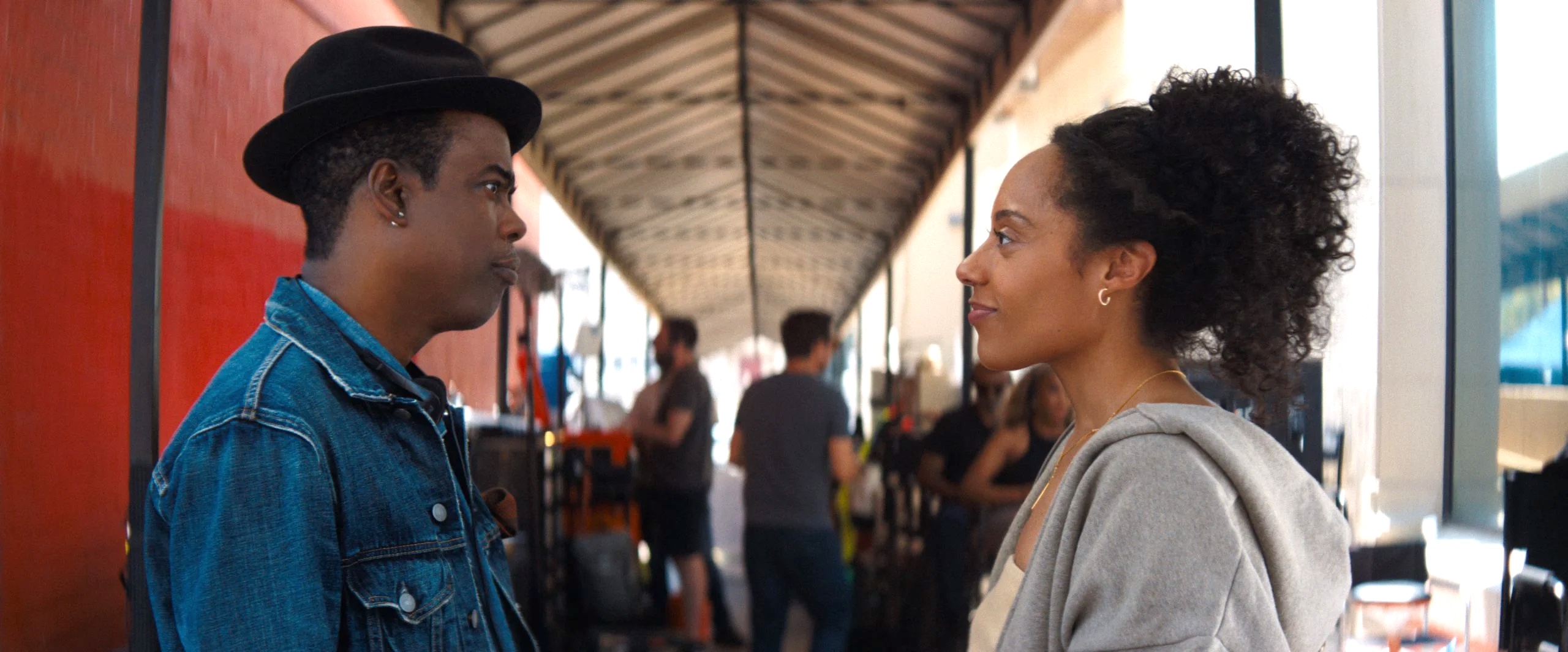 A man in a denim jacket and hat converses with a woman in a gray hoodie, both smiling, in a busy outdoor setting with people and equipment in the background.