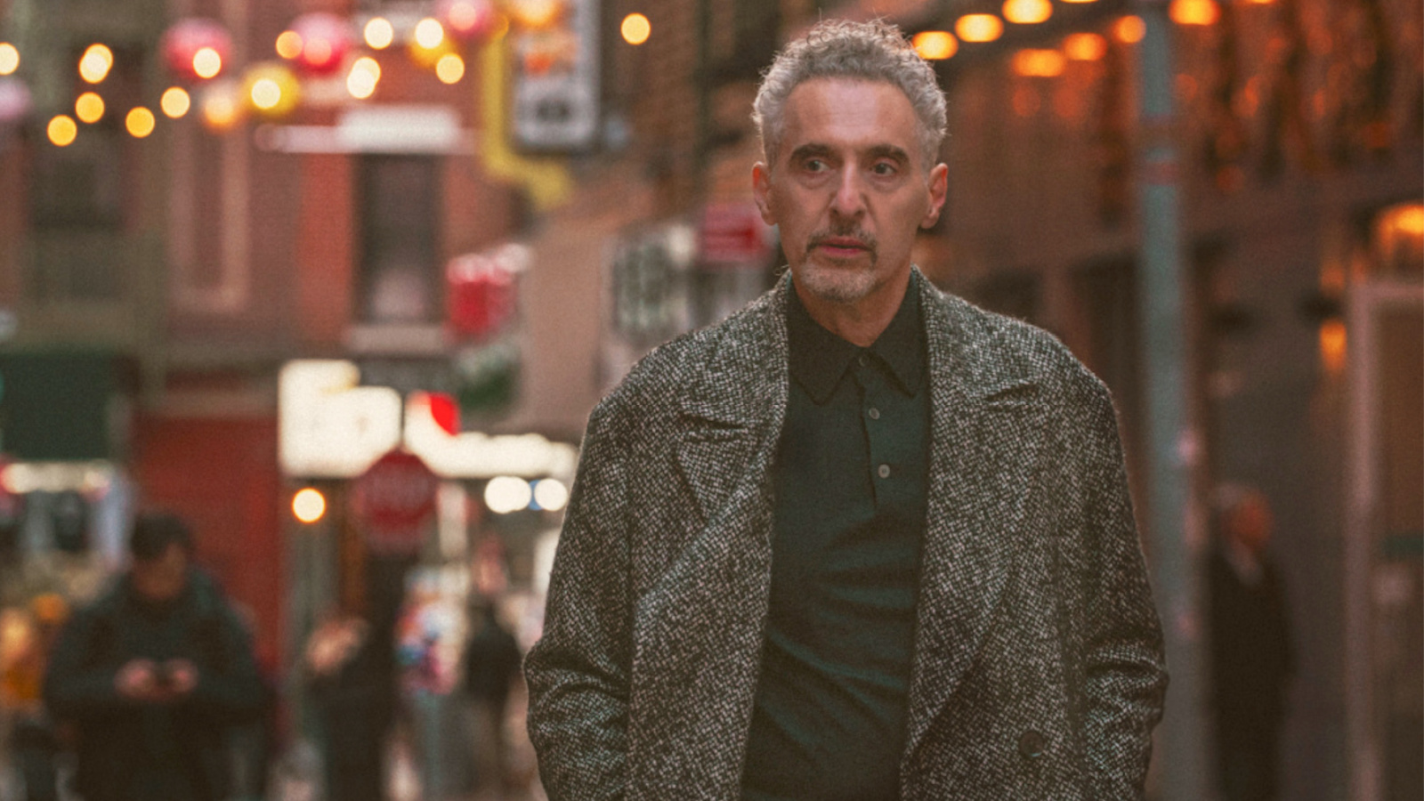 A man with gray hair and a beard walks confidently through a lively street adorned with colorful lights and signs.