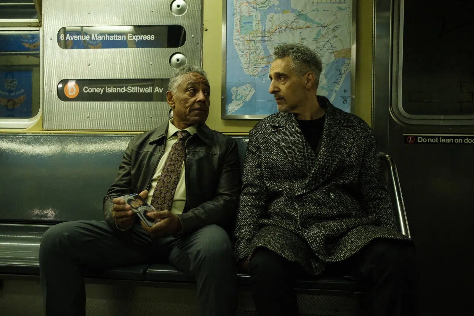 Two men sitting side by side on a subway bench, with one holding a photo. The subway interior features a map and route signs.
