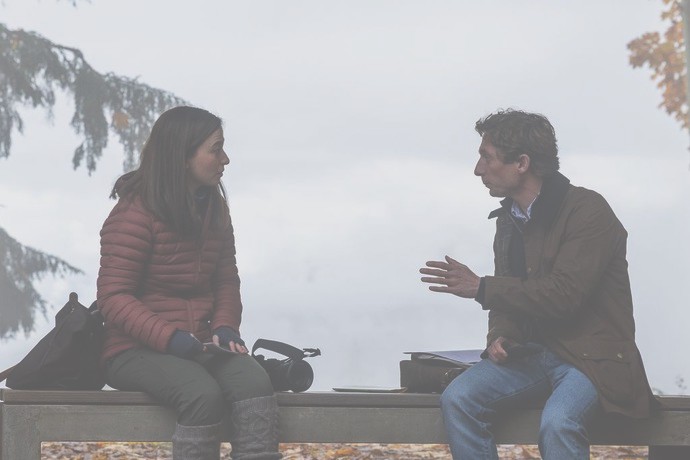 A man and a woman sit on a bench in a park, engaged in conversation, with autumn foliage in the background. The man gestures with his hand while speaking, and the woman listens attentively, both appearing thoughtful.